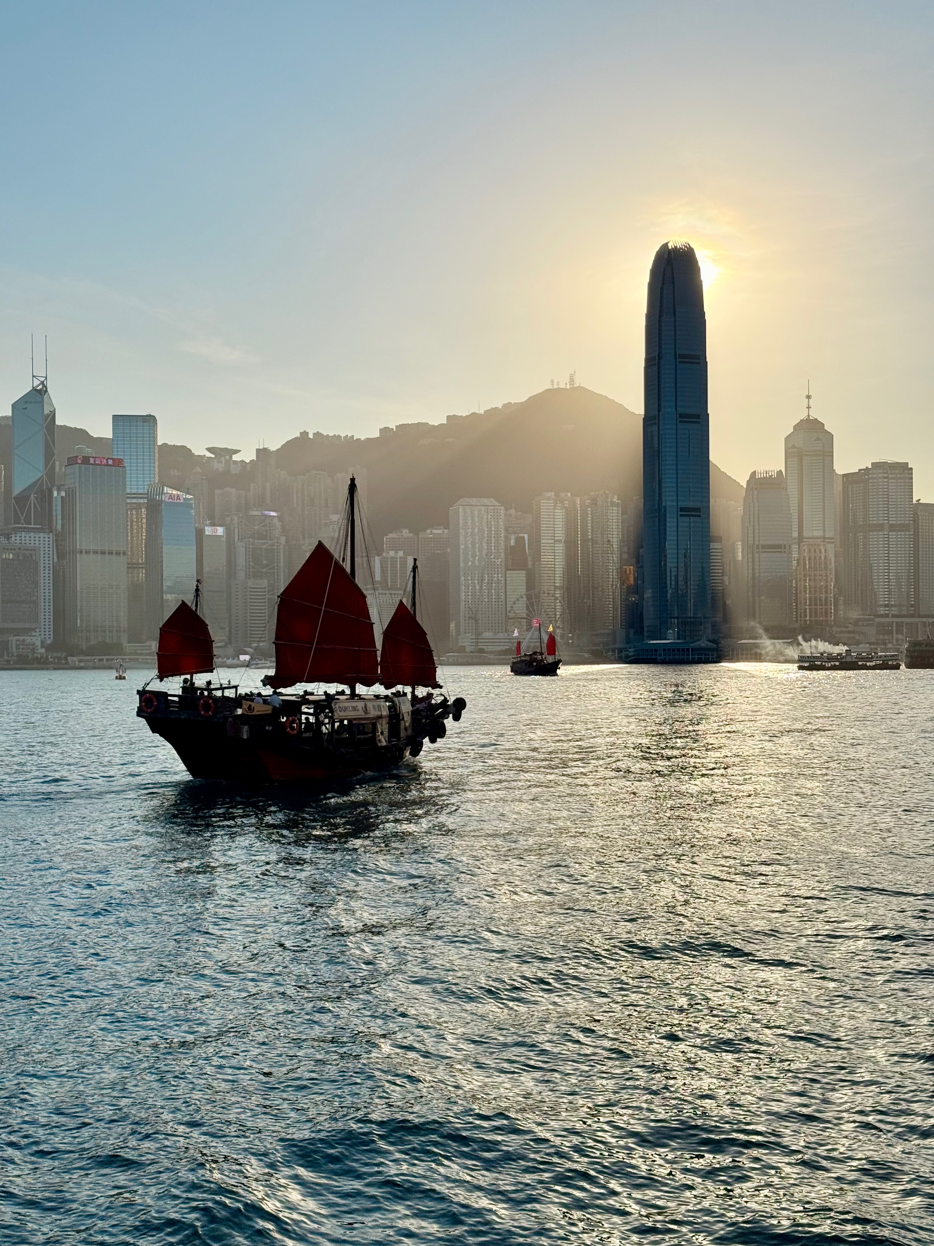 Photo of a Chinese junk with red sails on the water. There are hills and skyscrapers behind, with the famous IFC tower partially blocking the setting sun.