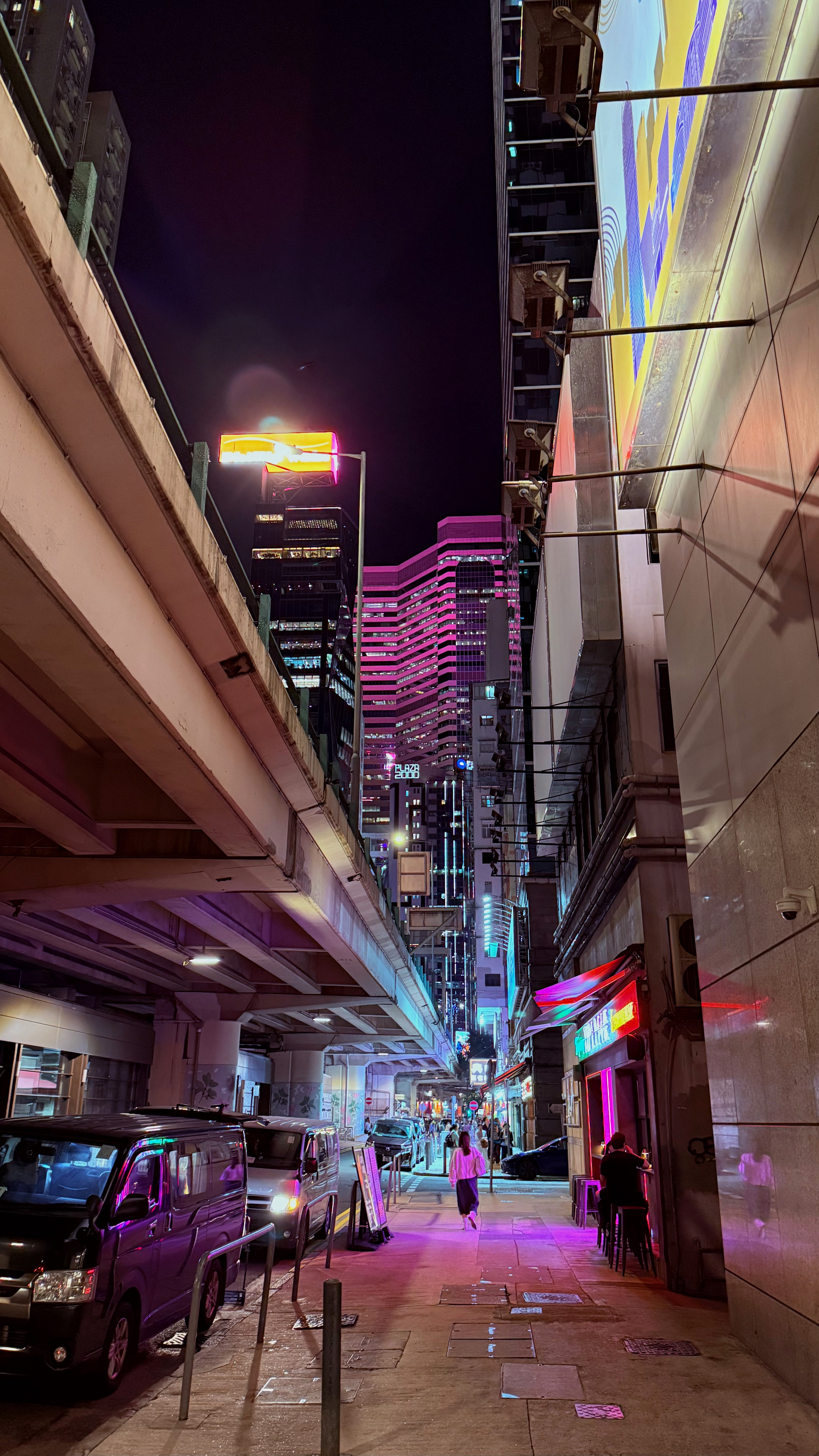 Wide angle photo of a street at night with an overpass on the screen left and a bar on the screen right. A woman walks through patches of magenta and blue light.