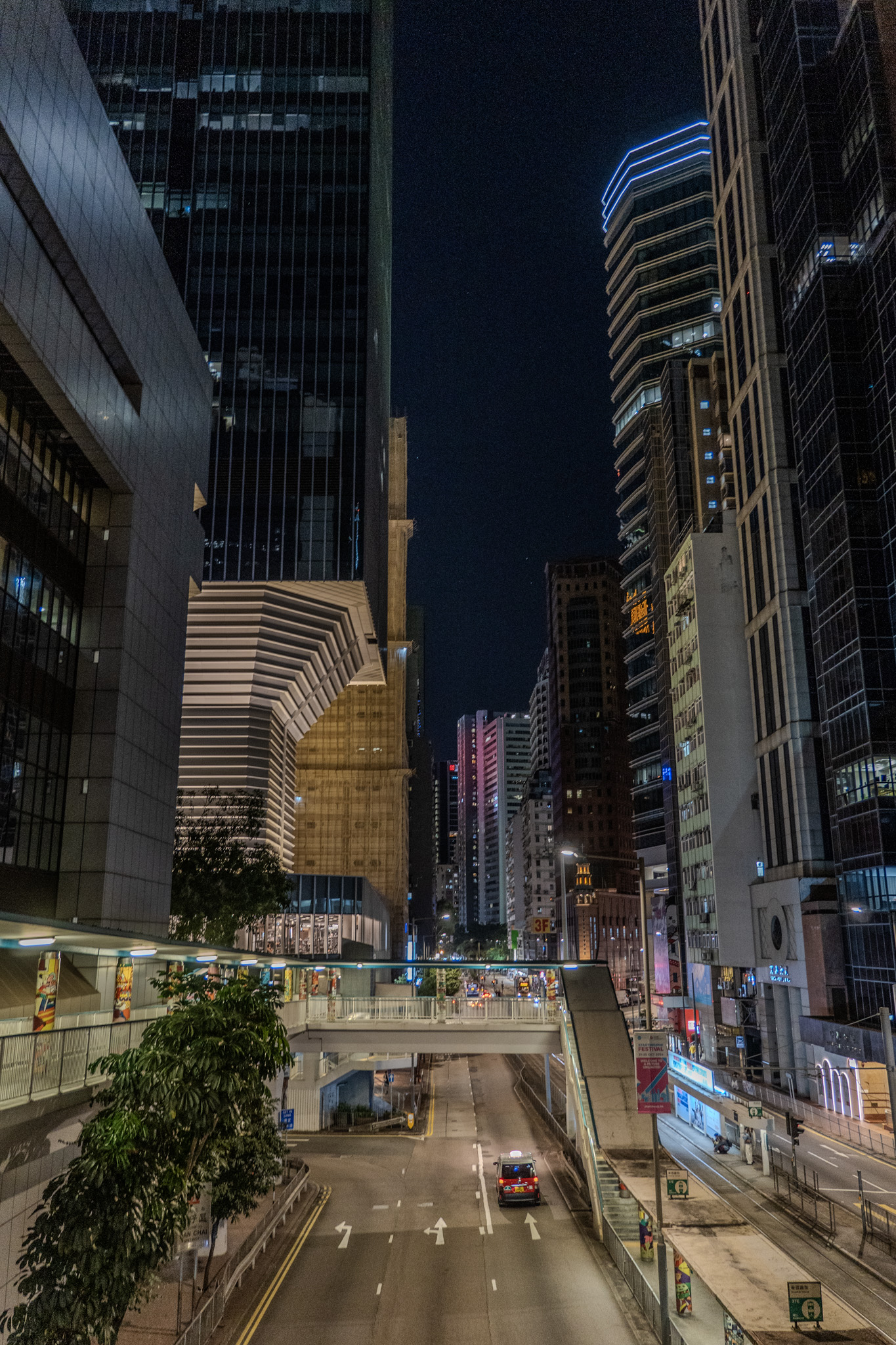 Wide angle photo of a street at night from above it on a pedestrian bridge. Part of the pedestrian bridge is on the screen left with people walking. The street below is empty except for one car. The building on the right is under construction with bamboo and green tarp skrims.