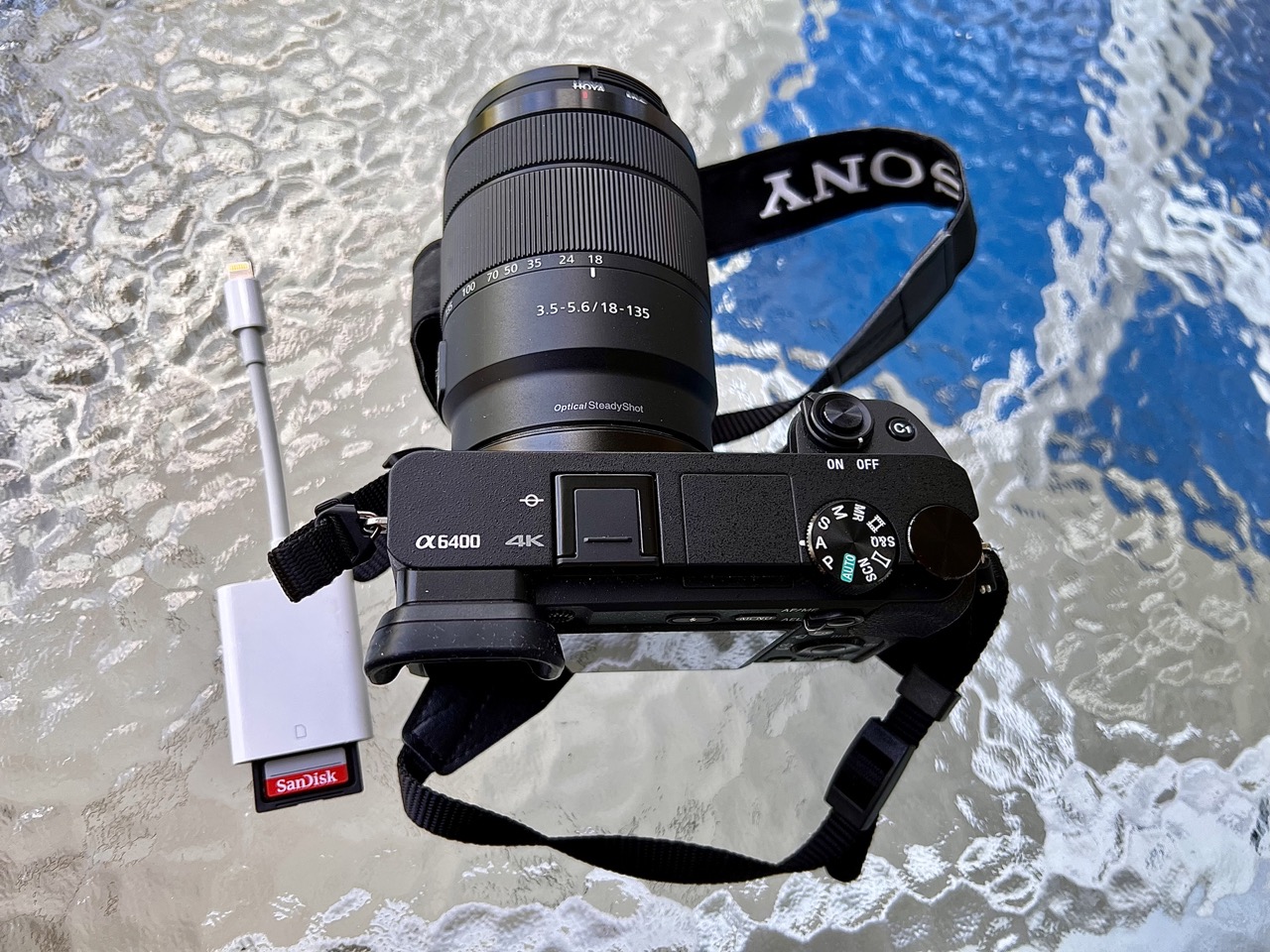 A photo of a Sony a6400 camera, and an Apple SD card reader, on top of one of those ugly rippled-glass patio tables.